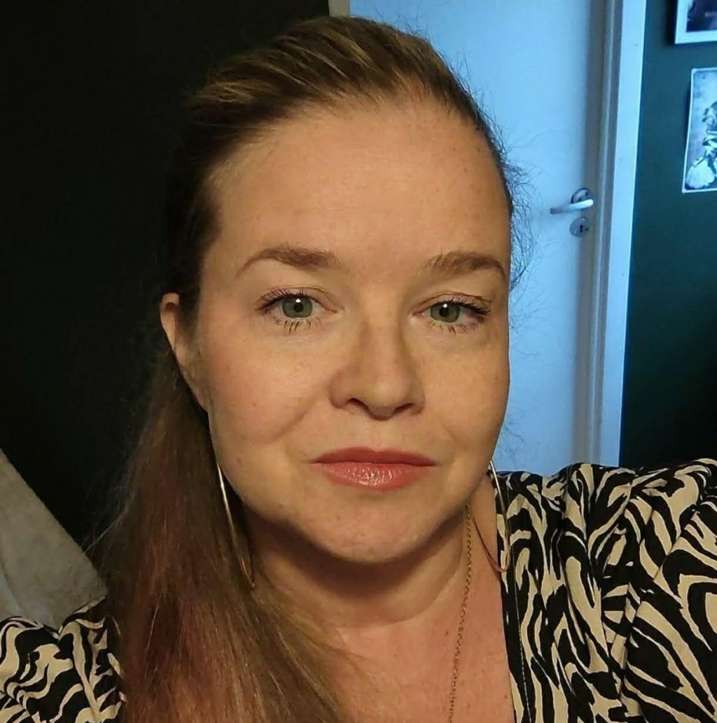 En kvinna med långt, ljusbrunt hår som är uppsatt, klädd i en mönstrad topp. Hon har ljus hy, är knappt sminkad och tittar rakt in i kameran. I bakgrunden syns en dörröppning och en del av en vägg med konstverk.