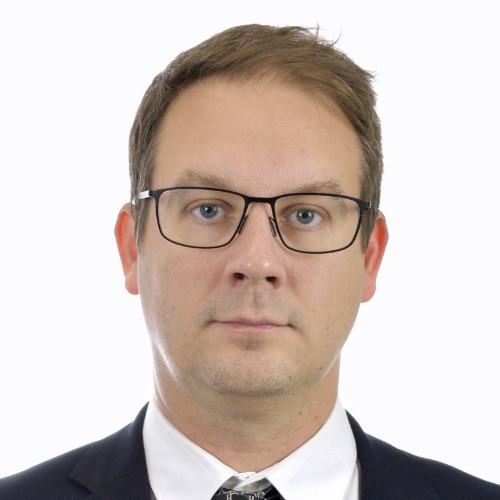 En man med kort ljusbrunt hår, som bär rektangulära glasögon, en svart kostymjacka, en vit skjorta och en mönstrad slips, poserar mot en enfärgad vit bakgrund och tittar rakt in i kameran med ett neutralt ansiktsuttryck.