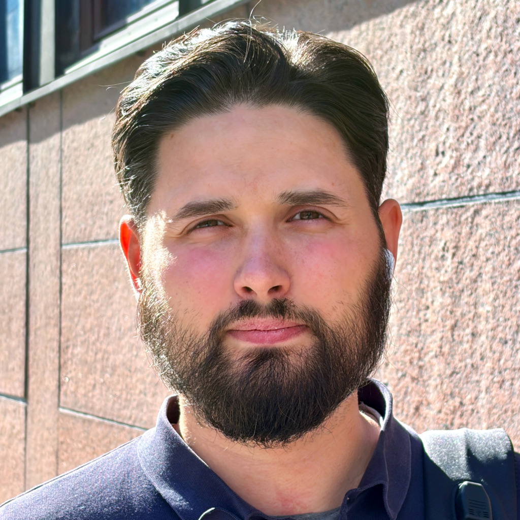 En man med mörkt hår och skägg står ute i solskenet framför en strukturerad stenvägg. Han har på sig en mörk skjorta med krage och man ser remmen till en väska på hans axel.