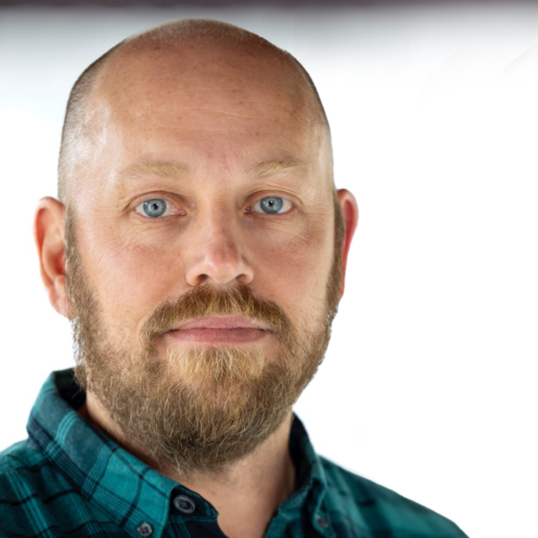 En man med rakat huvud, blå ögon och skägg bär en grön rutig skjorta och tittar rakt in i kameran mot en vit bakgrund.