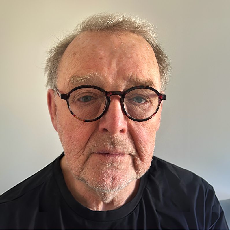 En äldre man med grått hår, som bär runda glasögon i sköldpaddsmönster och en svart skjorta, tittar rakt in i kameran. Bakgrunden är enfärgad och mjukt upplyst.