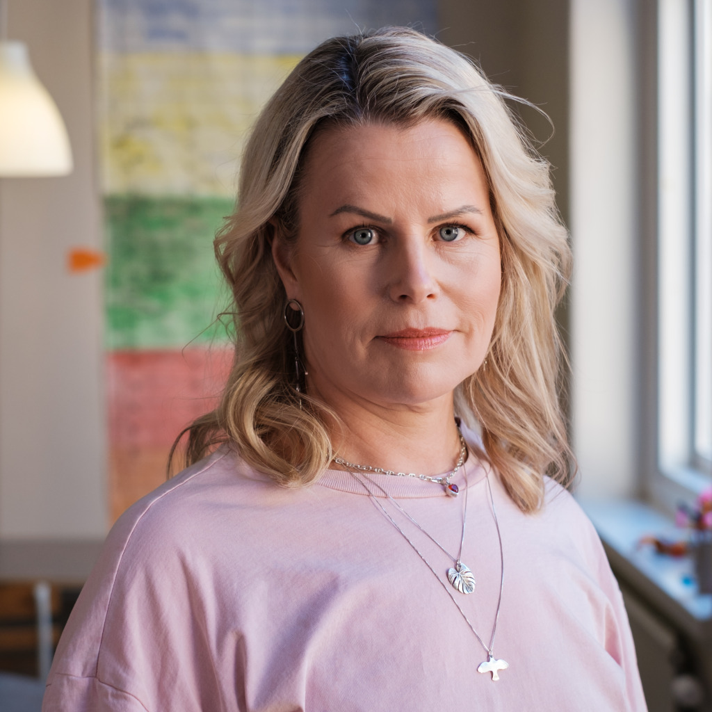 En kvinna med vågigt blont hår, klädd i en ljusrosa topp och flera halsband, står inomhus. Till höger om henne finns ett fönster och bakom henne syns en färgglad, suddig bakgrund. Belysningen är naturlig.