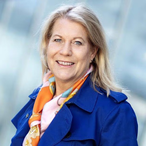 En blond kvinna ler mot kameran. Hon har på sig en blå kappa och en färgglad halsduk i orange och rosa nyanser. Bakgrunden är mjukt suddig med nyanser av blått och grått.