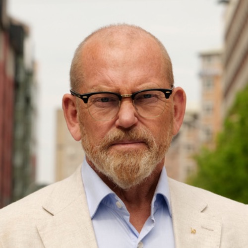 En medelålders man med skalligt huvud, rödaktigt skägg och glasögon har på sig en ljus kostymjacka och en blå skjorta. Han står utomhus på en gata i staden med byggnader och träd i bakgrunden.