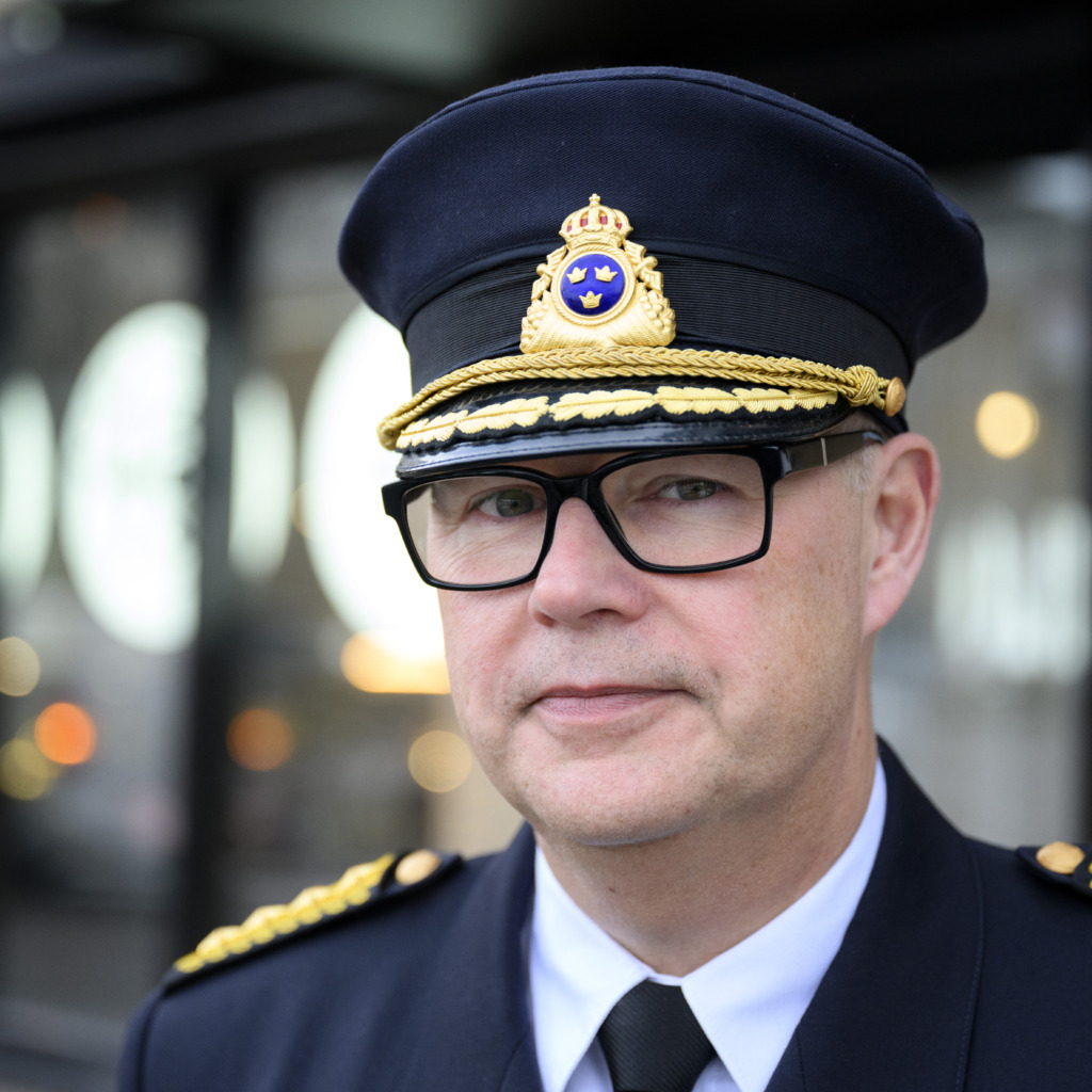 En man klädd i en mörk polisuniform, med en mössa med emblem, glasögon och ett allvarligt ansiktsuttryck står utomhus med suddiga ljus och byggnader i bakgrunden.