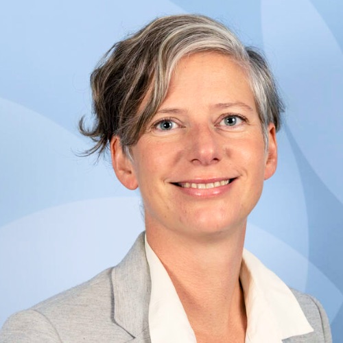 En person med kort, vågigt, brunt och grått hår ler. Hen bär en ljusgrå kavaj över en vit skjorta med krage – en klädsel som utstrålar professionalism och omsorg om arbetstiden. Bakgrunden är ljusblå med diskreta cirkulära mönster.