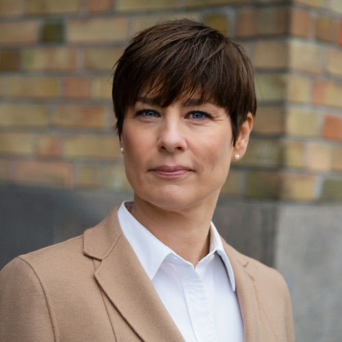 En person med kort brunt hår, klädd i en beige kavaj och en vit skjorta, står framför en suddig tegelvägg och tittar rakt in i kameran med ett neutralt ansiktsuttryck.