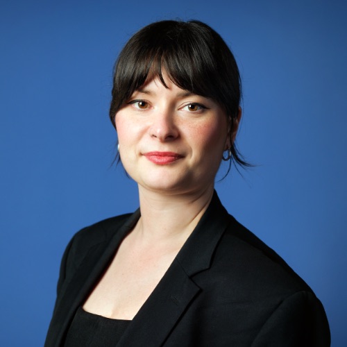 En kvinna med rakt, mörkt hår och lugg, klädd i en svart kavaj över en svart topp, poserar mot en enfärgad blå bakgrund med ett neutralt ansiktsuttryck.