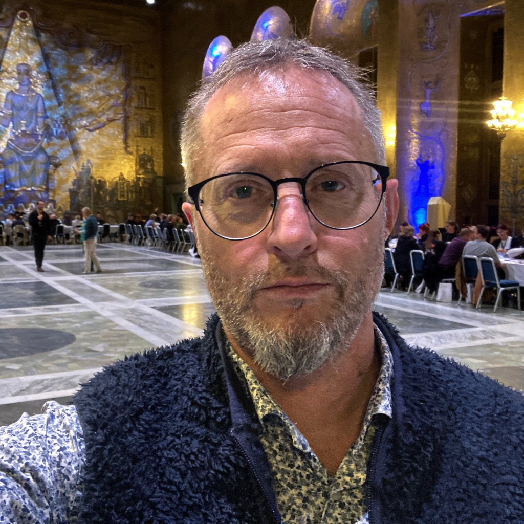 En man med glasögon och kort skägg tar en selfie i en storslagen sal med utsmyckade guldväggar, stora väggmålningar, välvda tak och matbord där människor sitter i bakgrunden.