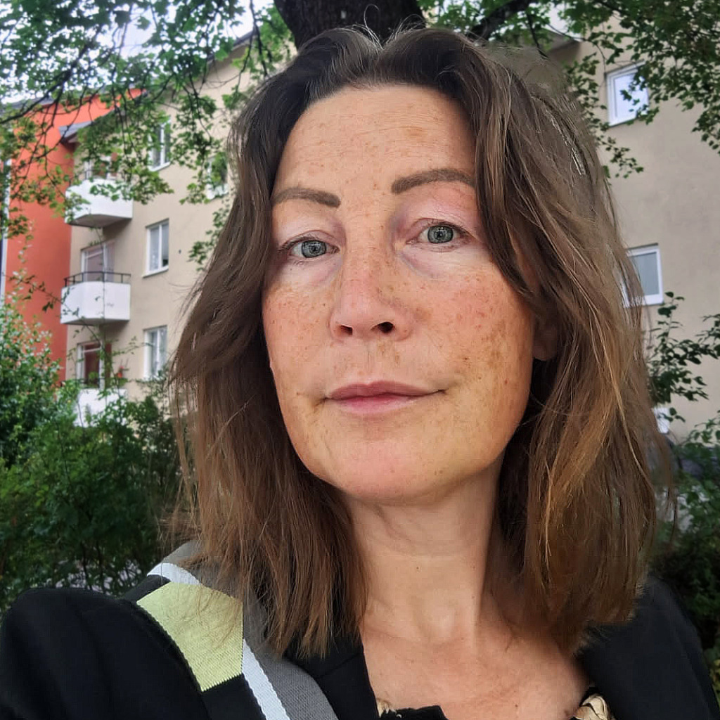 En person med axellångt brunt hår och ljus hy står utomhus framför ett flerbostadshus, med träd och grönska i bakgrunden. Personen tittar in i kameran med ett neutralt ansiktsuttryck.