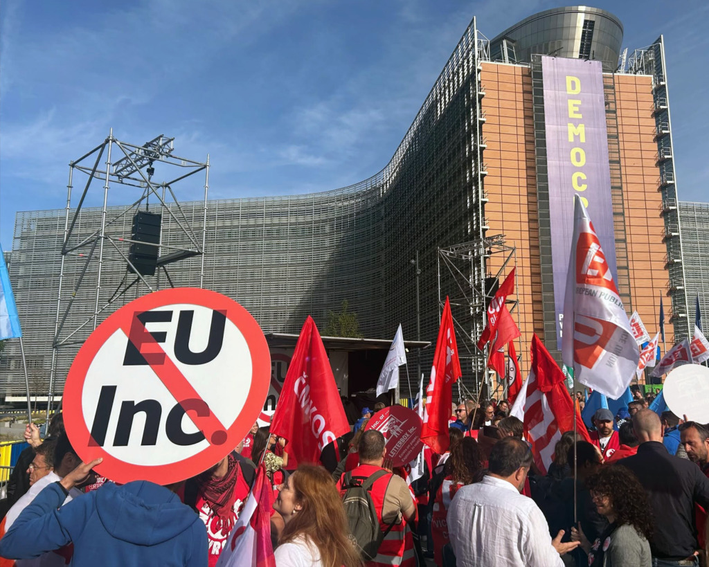 Demonstranter med plakat och flaggor samlas framför en stor EU-byggnad där en vertikal banderoll med texten ”DEMOKRATI” hänger.