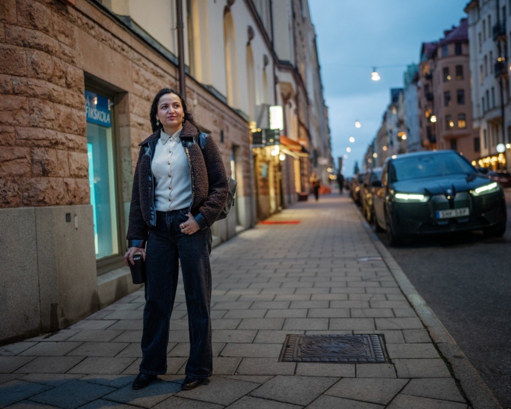 En kvinna står på en trottoar i stan i skymningen med en bil parkerad bredvid sig.