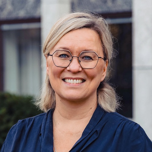 En leende kvinna med blont hår, mörka glasögon och en mörkblå skjorta.