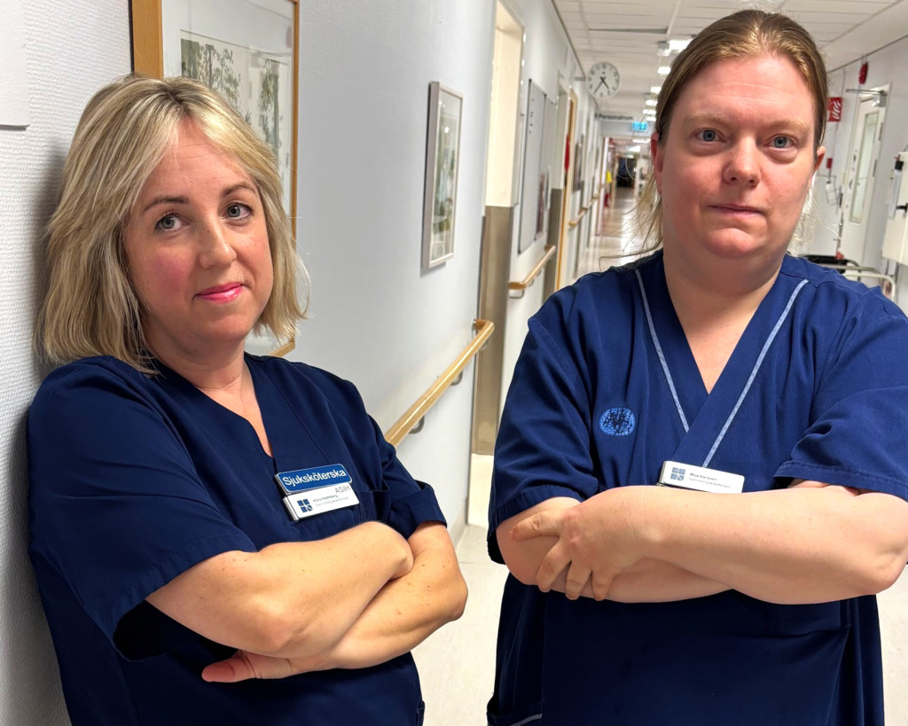 Två sjuksköterskor i mörkblå arbetskläder står i en sjukhuskorridor med armarna i kors.