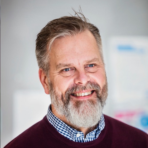 En man i medelåldern med grånande hår och skägg, klädd i en vinröd tröja över en blå- och vitrutig skjorta.