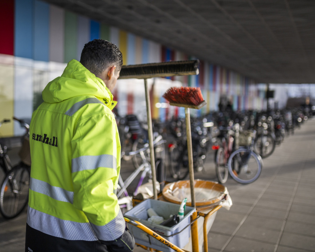En arbetare i en reflexväst skjuter en städvagn med kvastar genom en cykelparkering.