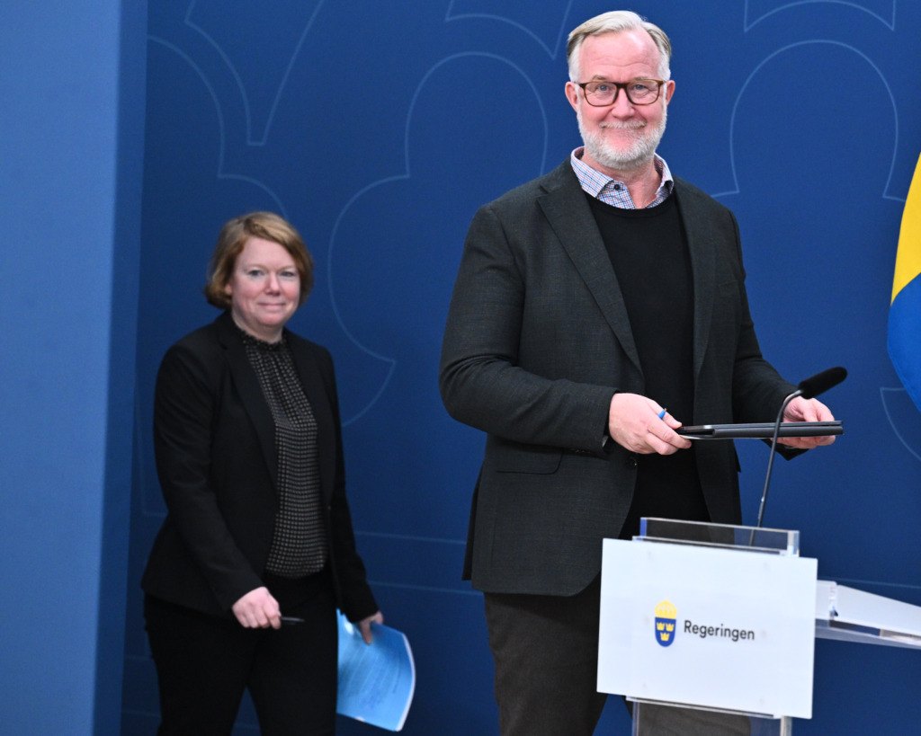 En man i kavaj står vid ett talarstativ med texten ”Regeringen”, medan en kvinna i kostym går bakom honom.