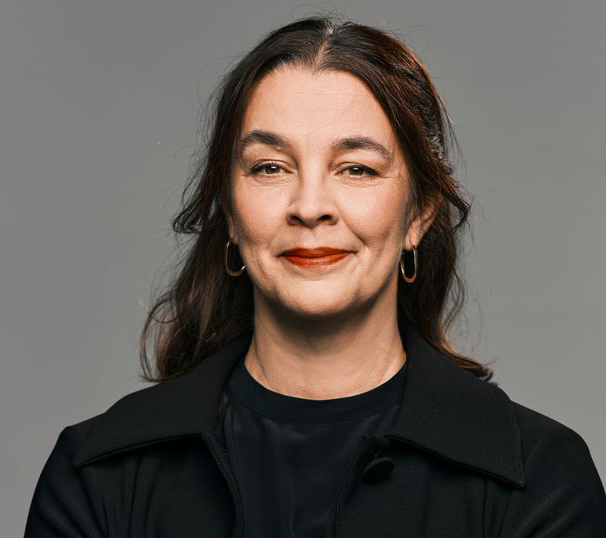 Sara Revell Ford, en medelålders kvinna med mörkt hår och guldfärgade ringörhängen, klädd i en svart jacka mot en grå bakgrund.